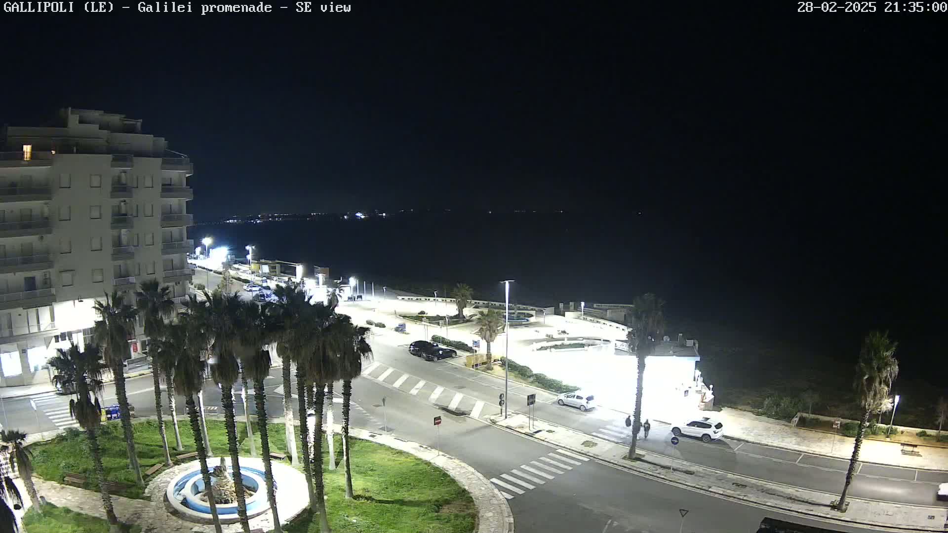 A nighttime, high-angle view shows a seaside promenade with palm trees, a building, cars, and a fountain in a small roundabout.