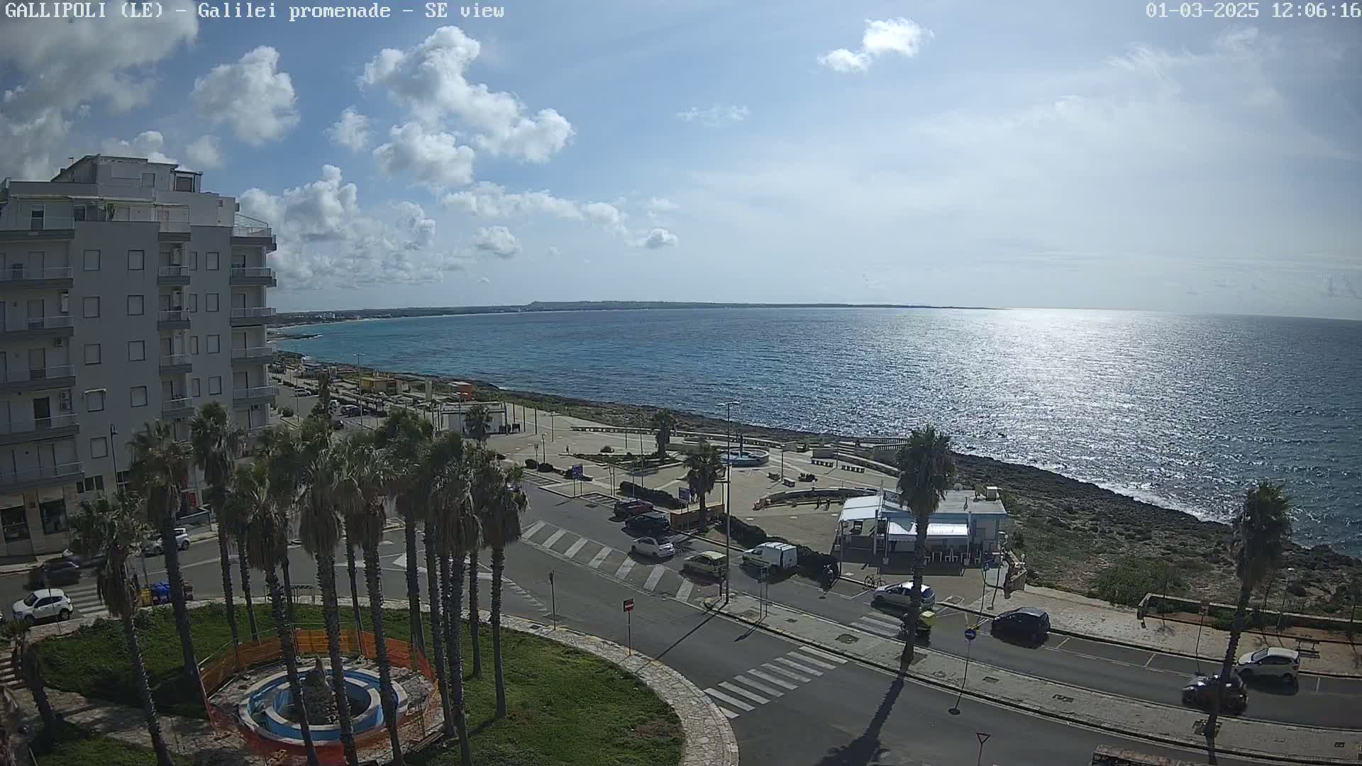 A partly sunny day overlooking a calm sea from a coastal town with palm trees lining a roadway next to a building.