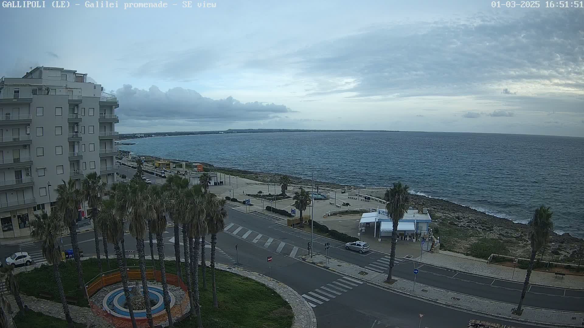 An overcast day reveals a coastal town scene with a calm sea, palm trees lining a road that curves toward a seaside promenade.