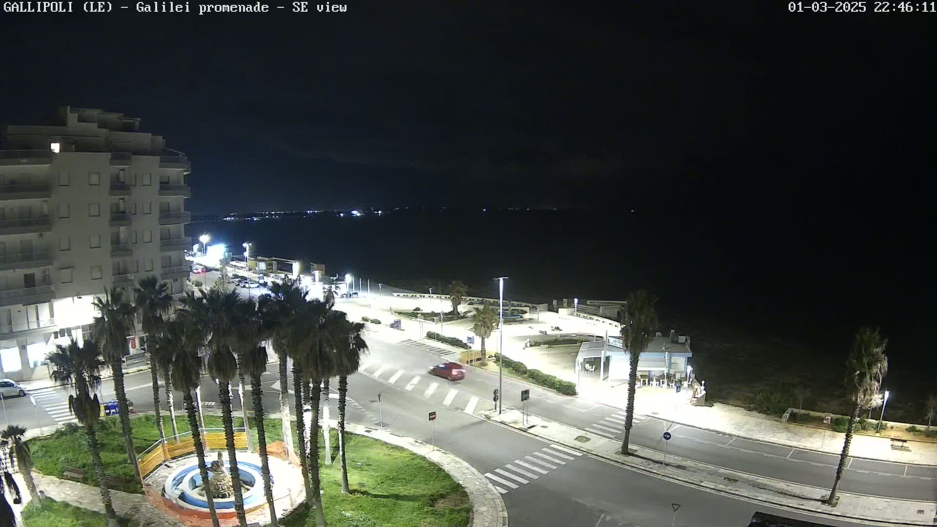 A nighttime, high-angle view of a seaside town with a multi-story building, palm trees lining a roadway, and a dark body of water visible in the distance.