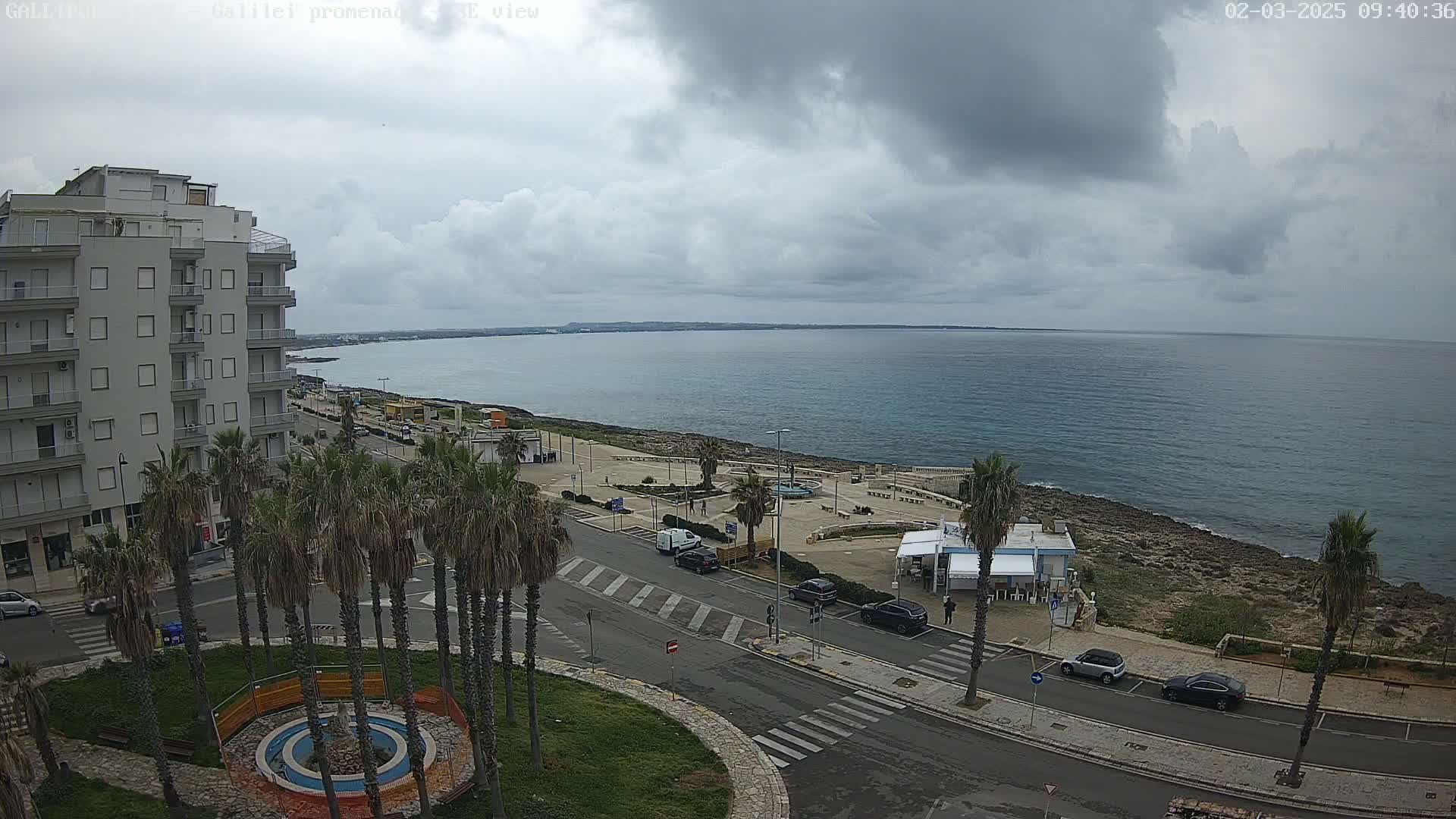 A coastal town scene under a cloudy sky shows a multi-story building, palm trees lining a street, a waterfront promenade, and a calm sea extending to the horizon.