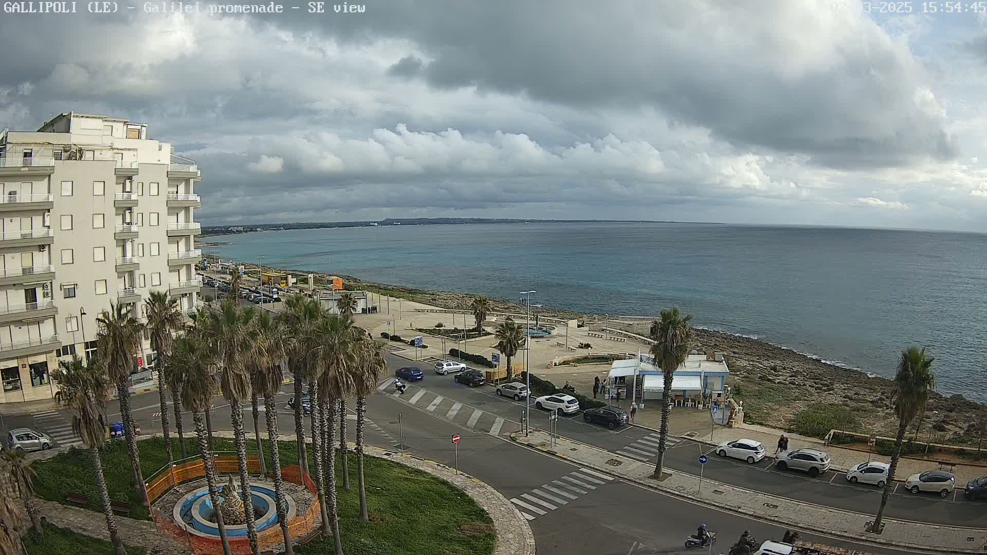 A coastal scene under a mostly cloudy sky shows a multi-story building, palm trees, parked cars, a roadway, and a calm sea extending to a distant horizon.