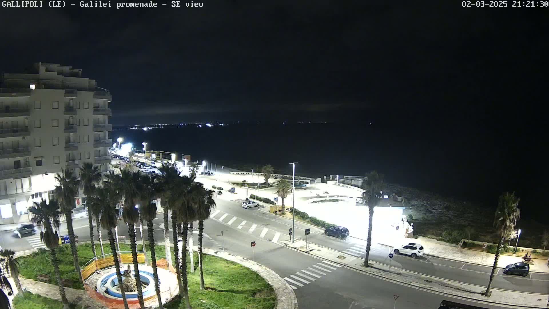 A nighttime, high-angle view of a coastal town's promenade shows a mostly empty parking lot, a few cars on the road, and palm trees lining a circular area with a fountain.
