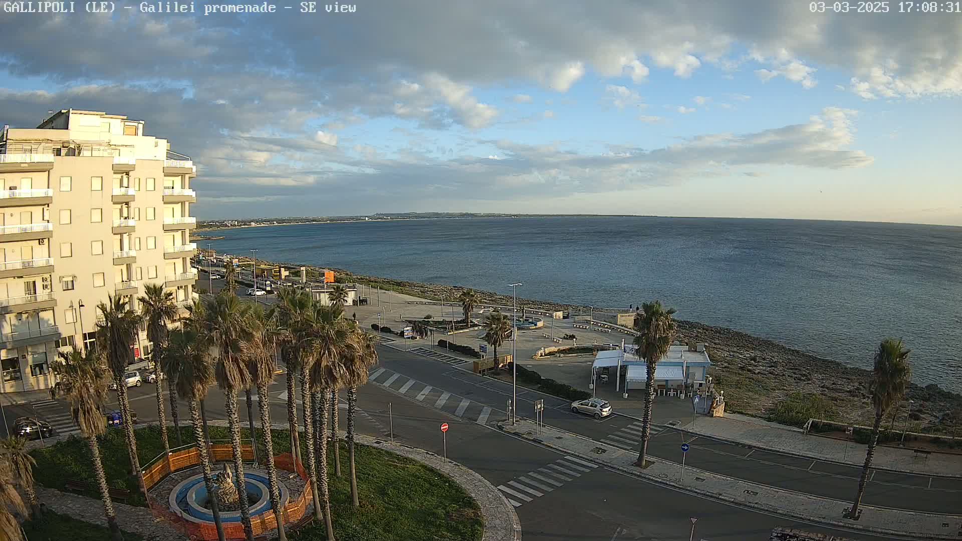 An aerial view of a seaside promenade on a partly cloudy day shows a calm sea, palm trees, a multi-story building, and some parked cars.