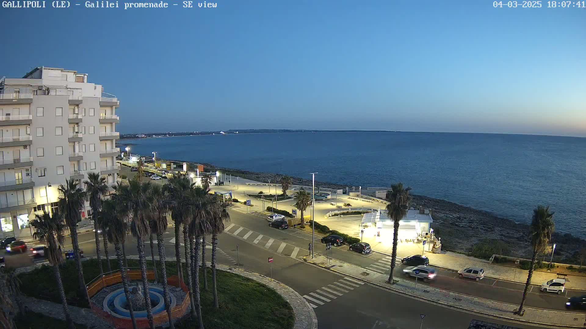A seaside town at dusk, with palm trees lining a street next to a calm, dark blue sea.