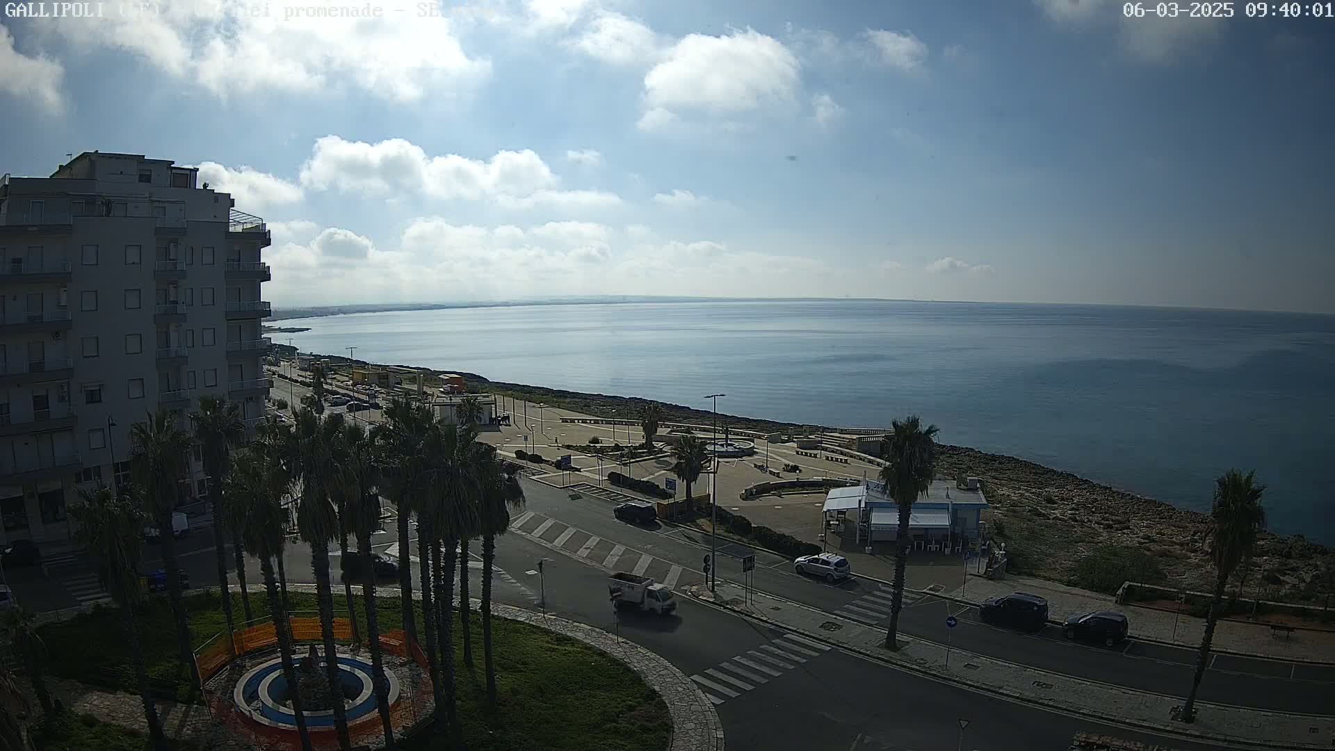 An elevated view shows a partly cloudy sky over a calm sea, a coastal road with parked cars, palm trees, and a multi-story building.