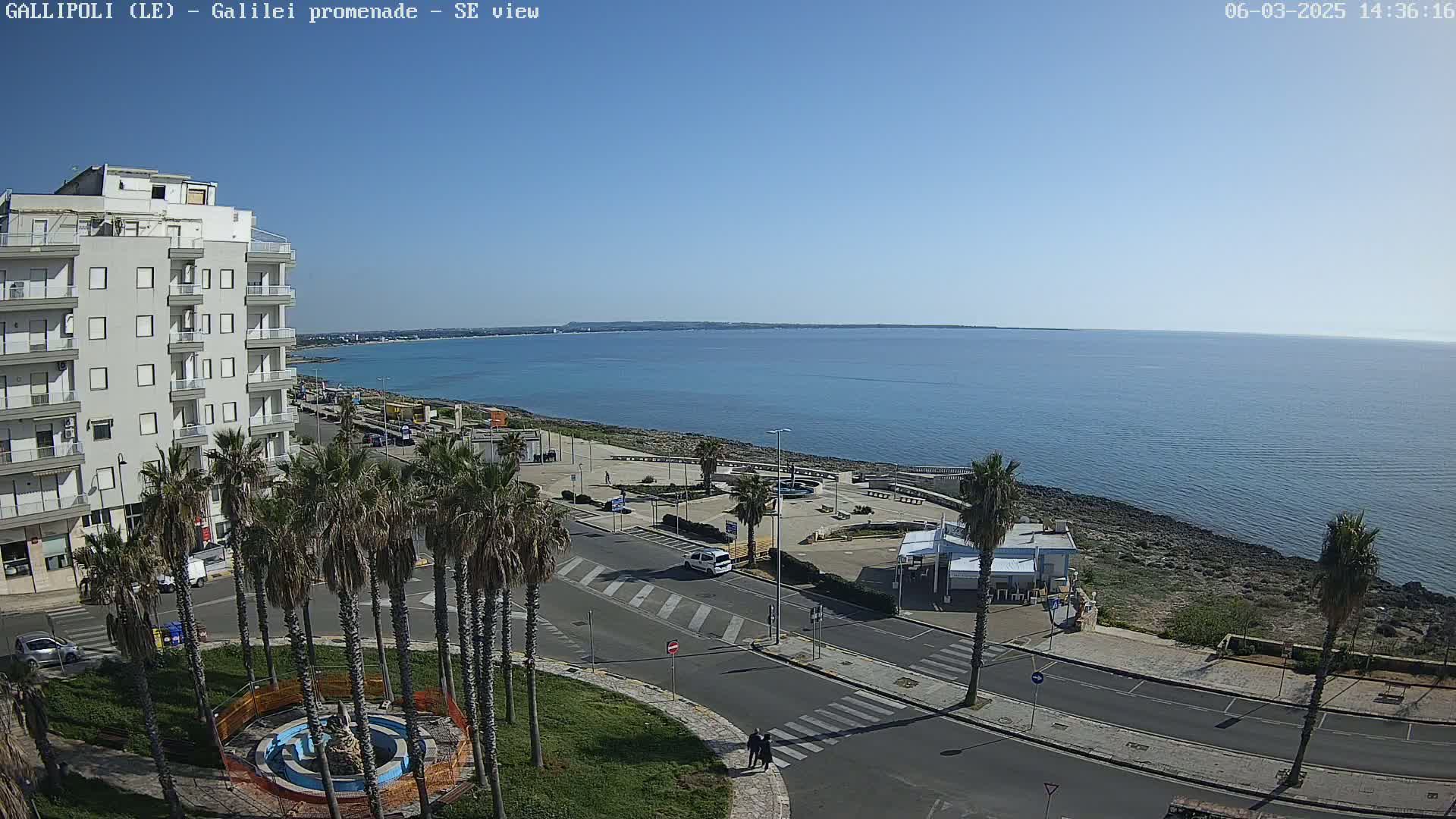 A seaside town scene on a clear, sunny day shows a multi-story building, palm trees lining a road, a rocky shoreline, and a calm blue sea extending to the horizon.