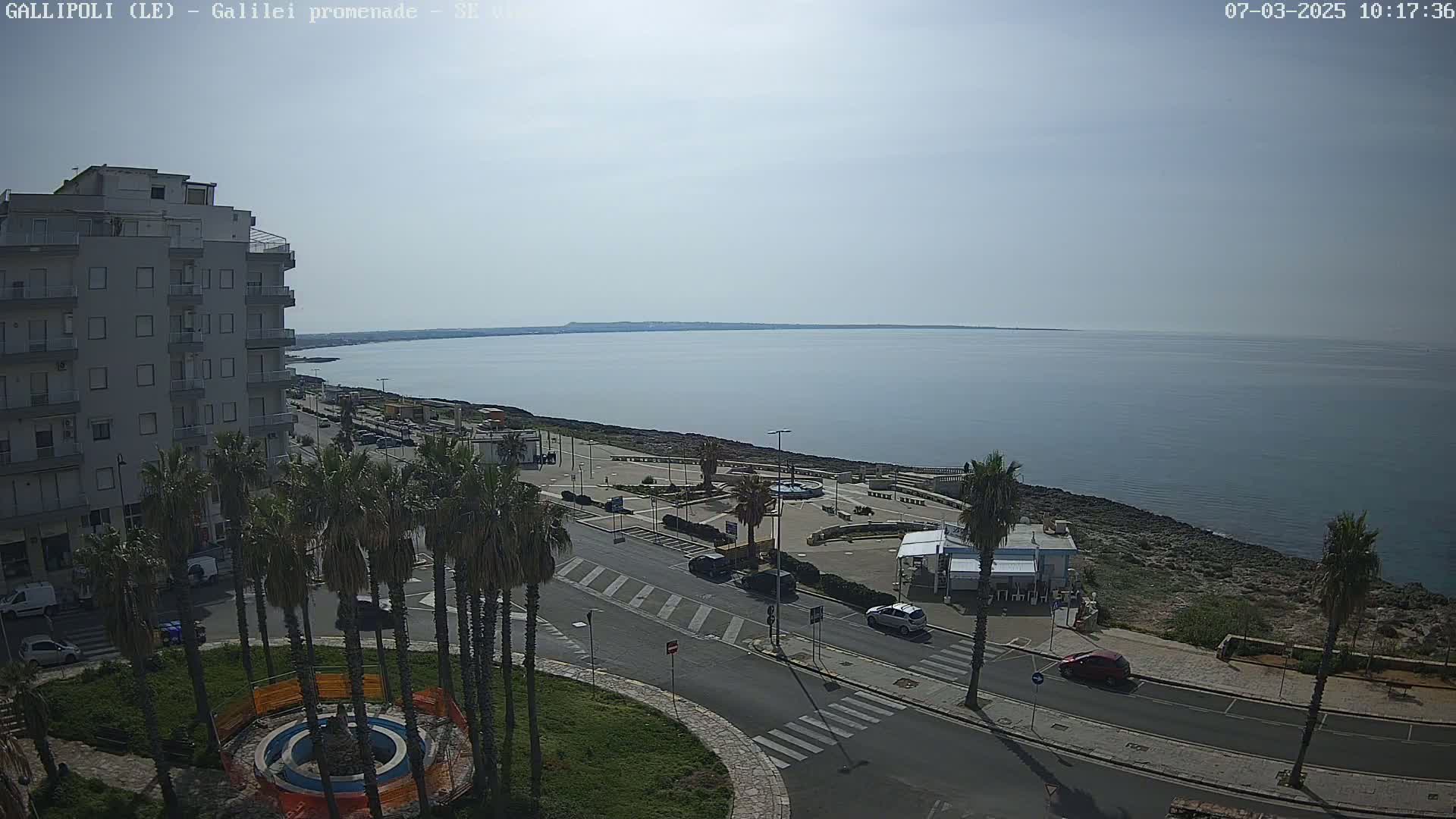 A mostly sunny day overlooking a calm sea from a coastal town with palm trees, a multi-story building, and a paved road.