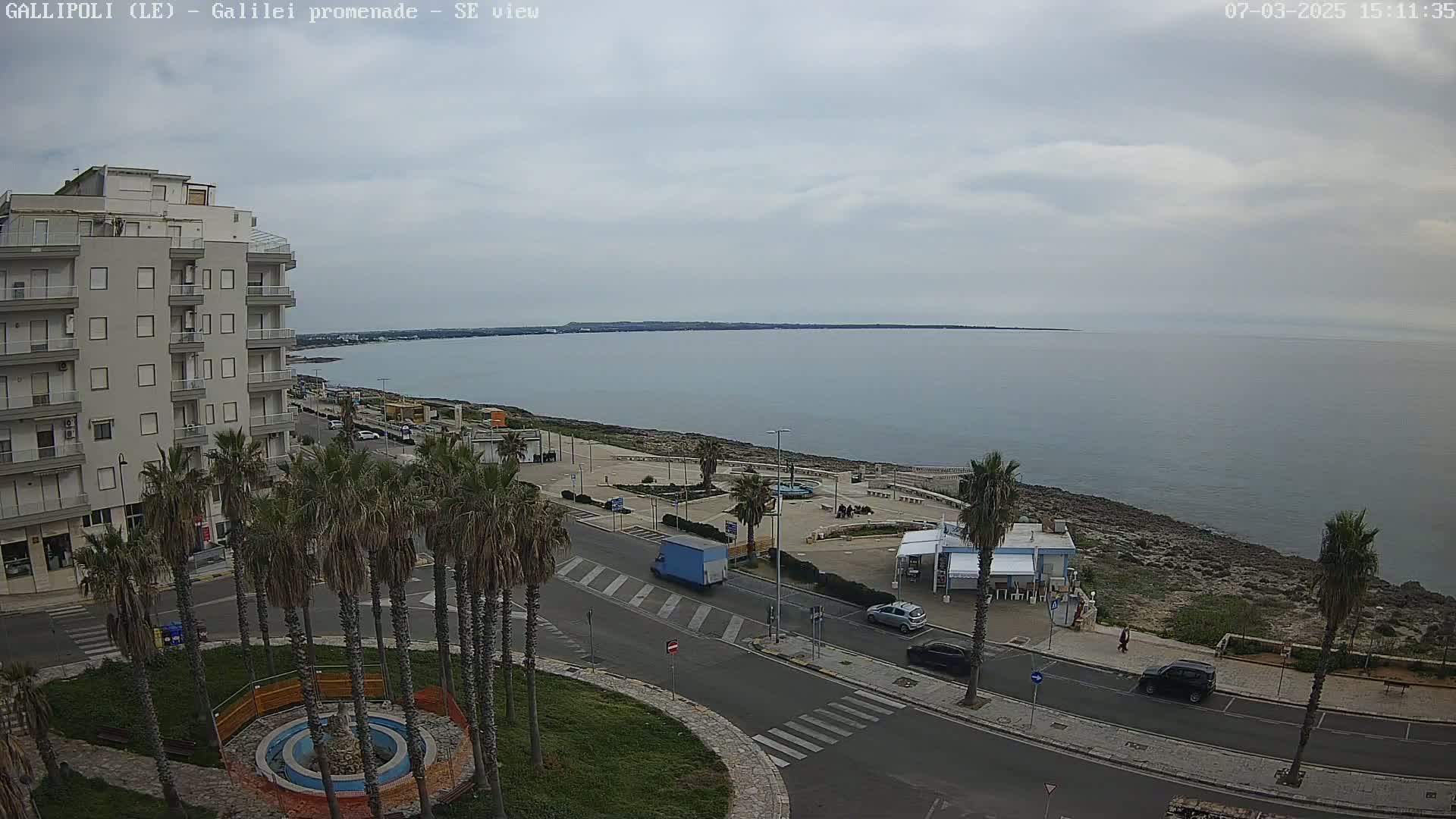An overcast day reveals a seaside town with palm trees lining a road next to a calm sea, and a multi-story building visible in the foreground.