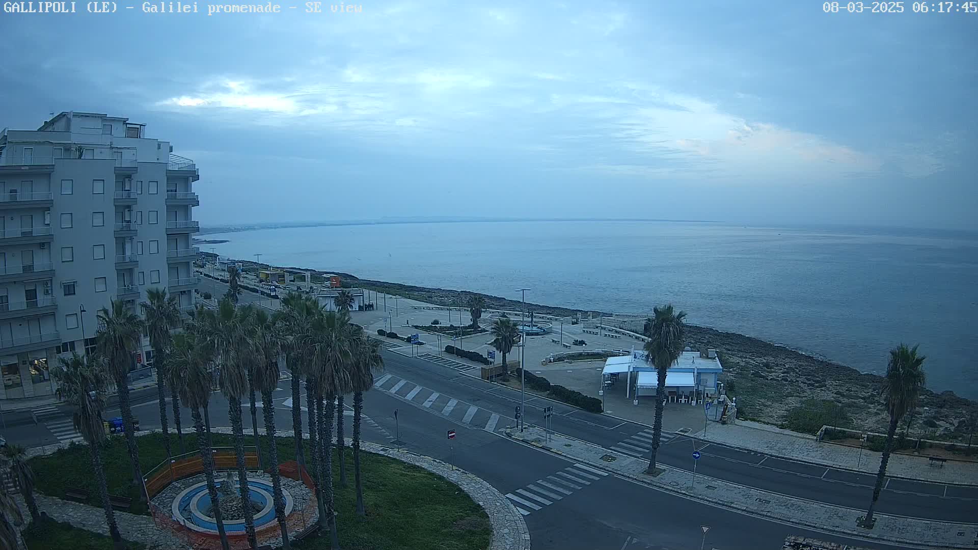 A mostly cloudy morning reveals a coastal town scene with a calm sea, palm trees lining a road next to a promenade overlooking the water, and a multi-story building in the foreground.