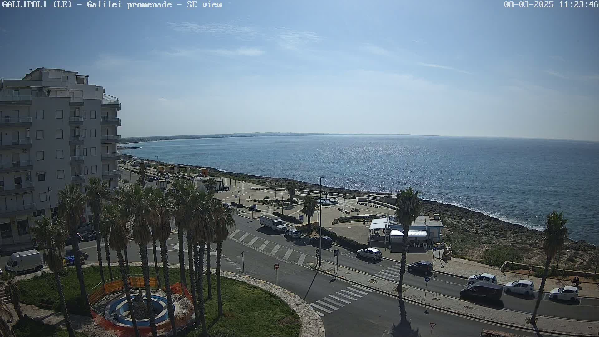 A seaside town on a sunny day shows a multi-story building, palm trees, a promenade along the coast, and cars parked on a street overlooking the ocean.