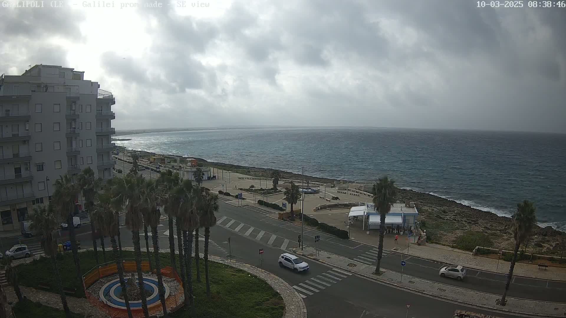 A seaside town under a cloudy sky shows a multi-story building, palm trees, a roadway, and a rocky coastline with a calm sea beyond.