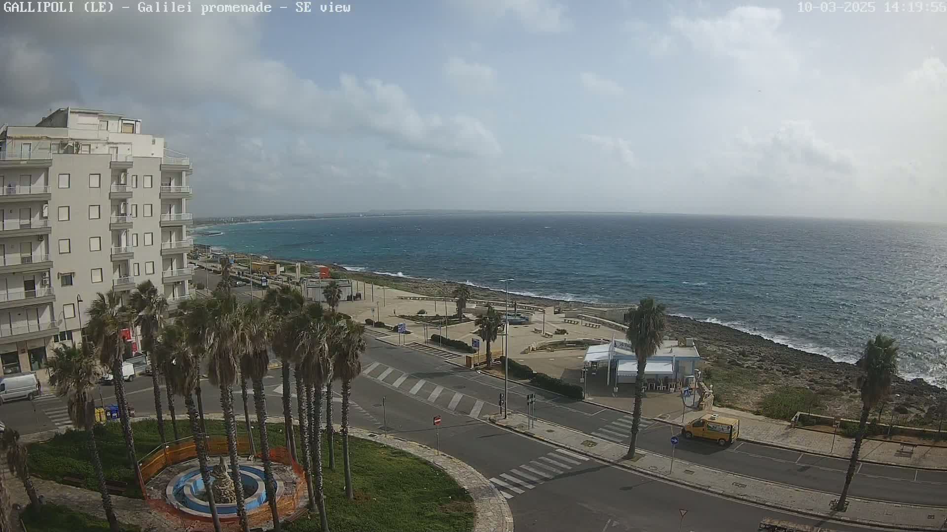 A partly cloudy day overlooking a seaside town with palm trees lining a road next to a calm, blue ocean.