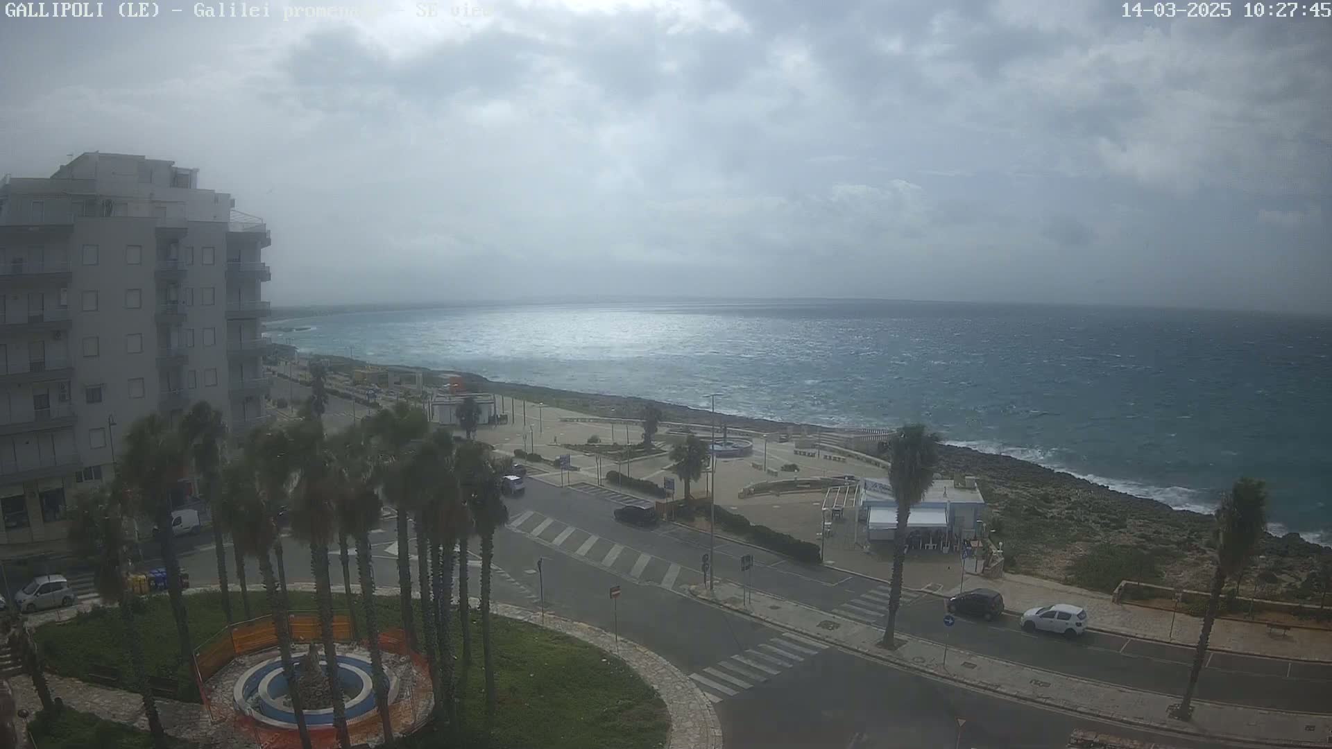 A coastal town scene under a cloudy sky shows a multi-story building, palm trees, a road, and a partly cloudy sky overlooking a choppy sea.