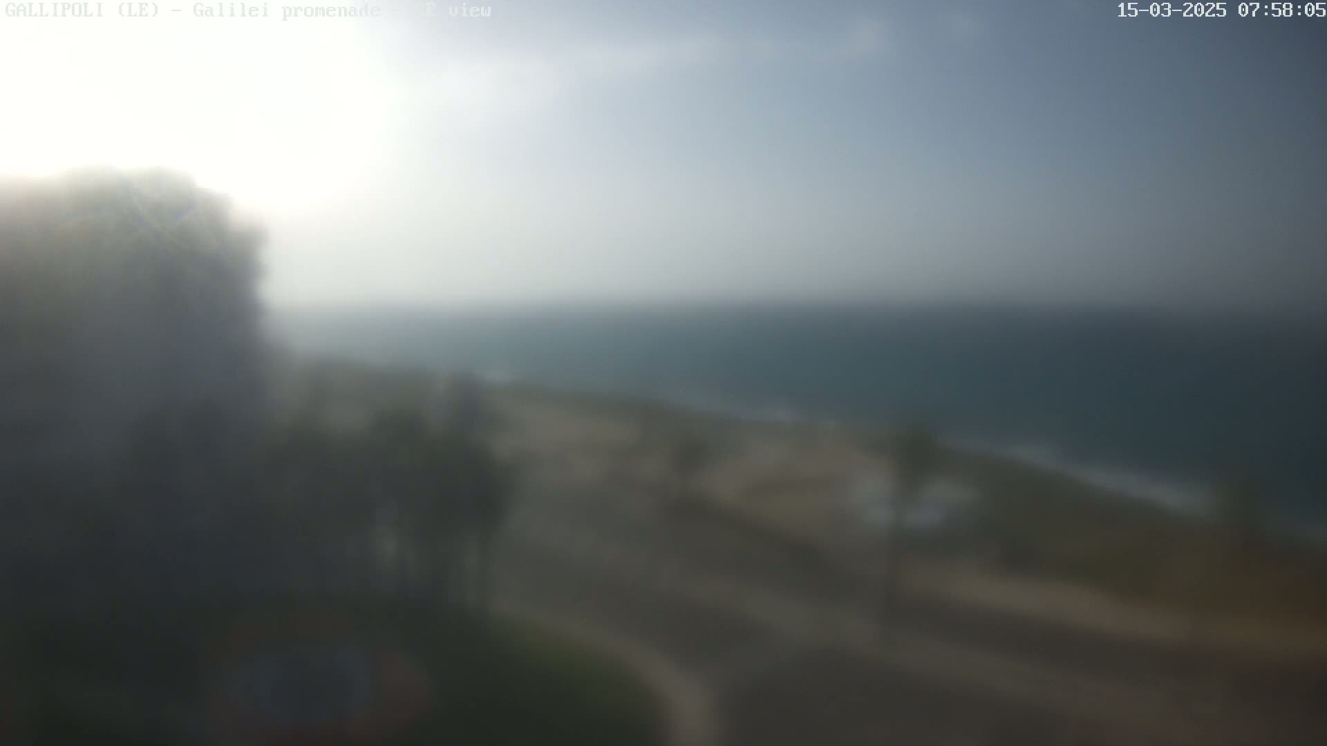 A hazy, slightly overcast view shows a coastline with a calm sea, vegetation, and a roadway.