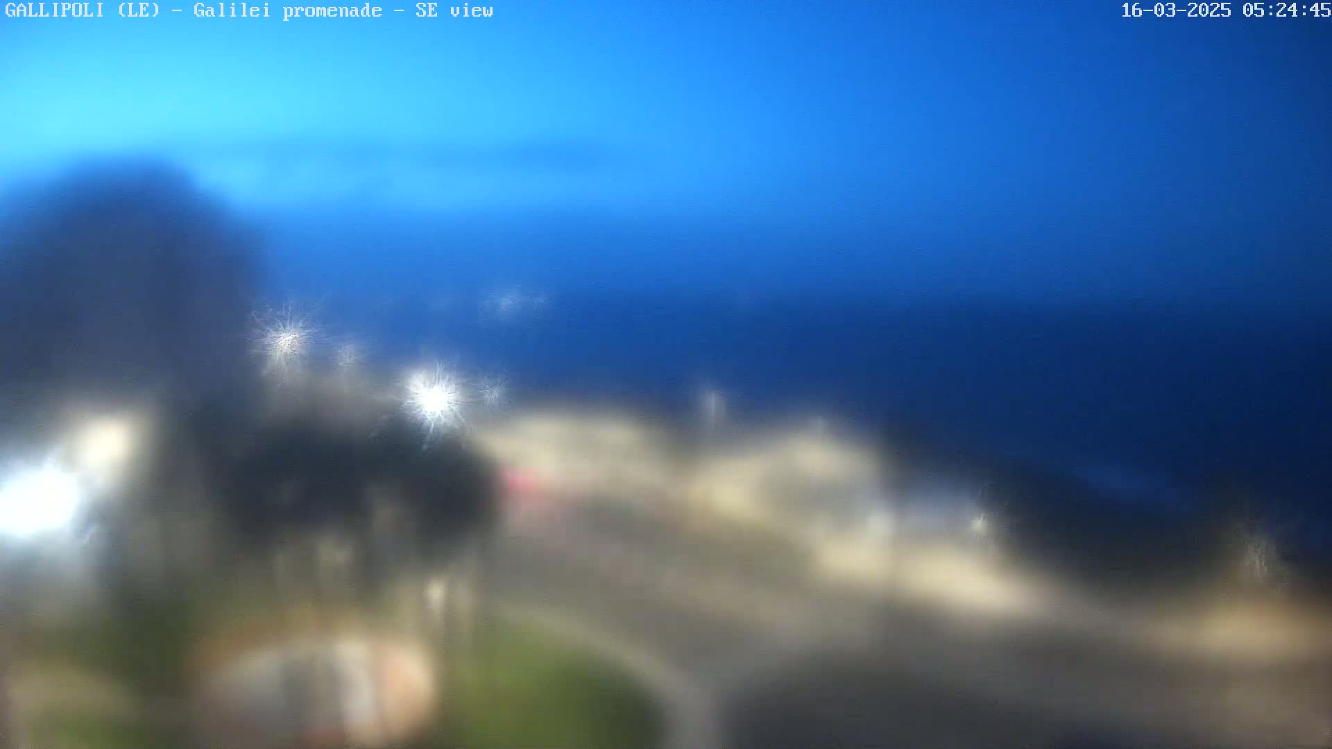 A blurry, nighttime view of a coastal town under a dark blue sky shows lights from buildings and streets near the water.