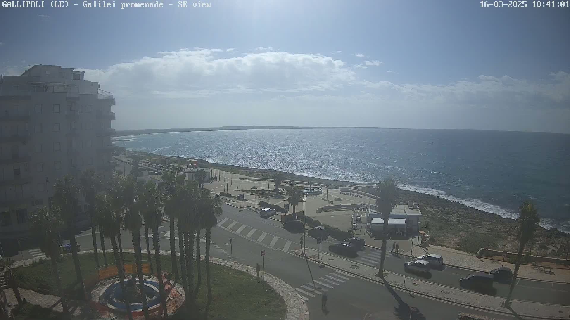 A partly cloudy day overlooking a coastal town with palm trees, a roadway, and a calm ocean.