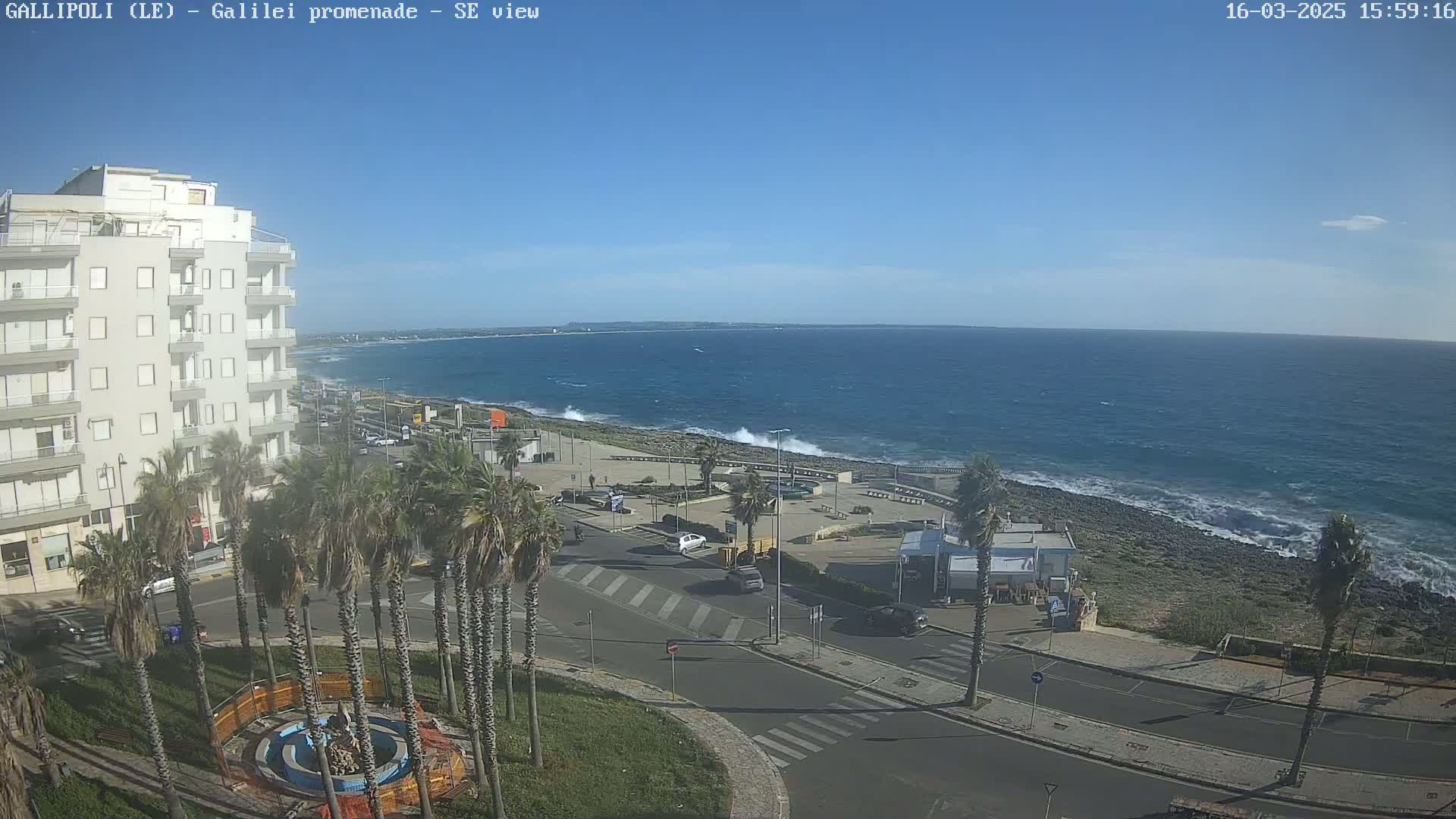 A seaside town with palm trees, a multi-story building, and a paved road overlooking a rocky shoreline with moderate waves under a clear blue sky.