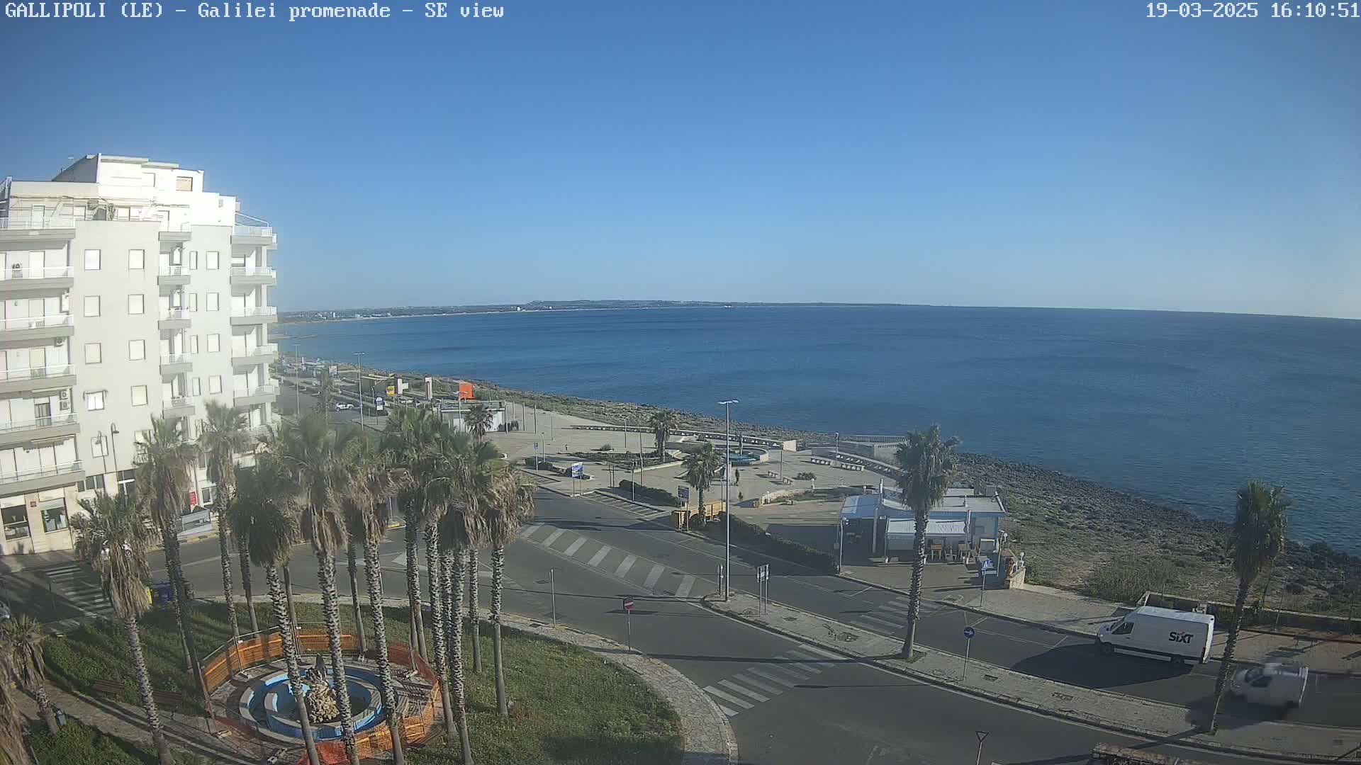 A coastal city scene on a sunny day shows a multi-story building, palm trees lining a road, a waterfront promenade, and a calm blue sea extending to the horizon.