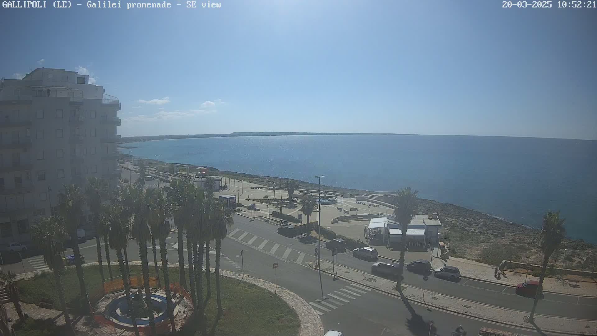 A seaside town scene on a sunny day shows a calm blue ocean, a paved promenade with parked cars, palm trees, and buildings, a small roundabout with a fountain in the foreground.