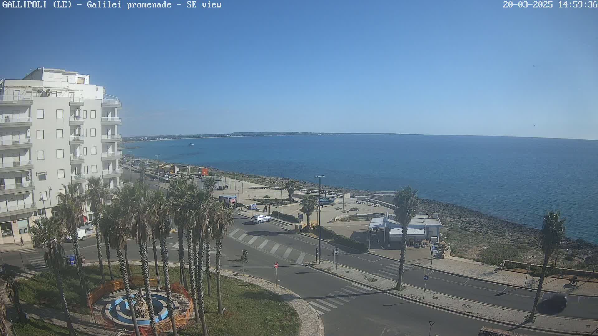 A seaside town on a sunny day shows a calm blue sea, a paved road with palm trees, and a multi-story building.