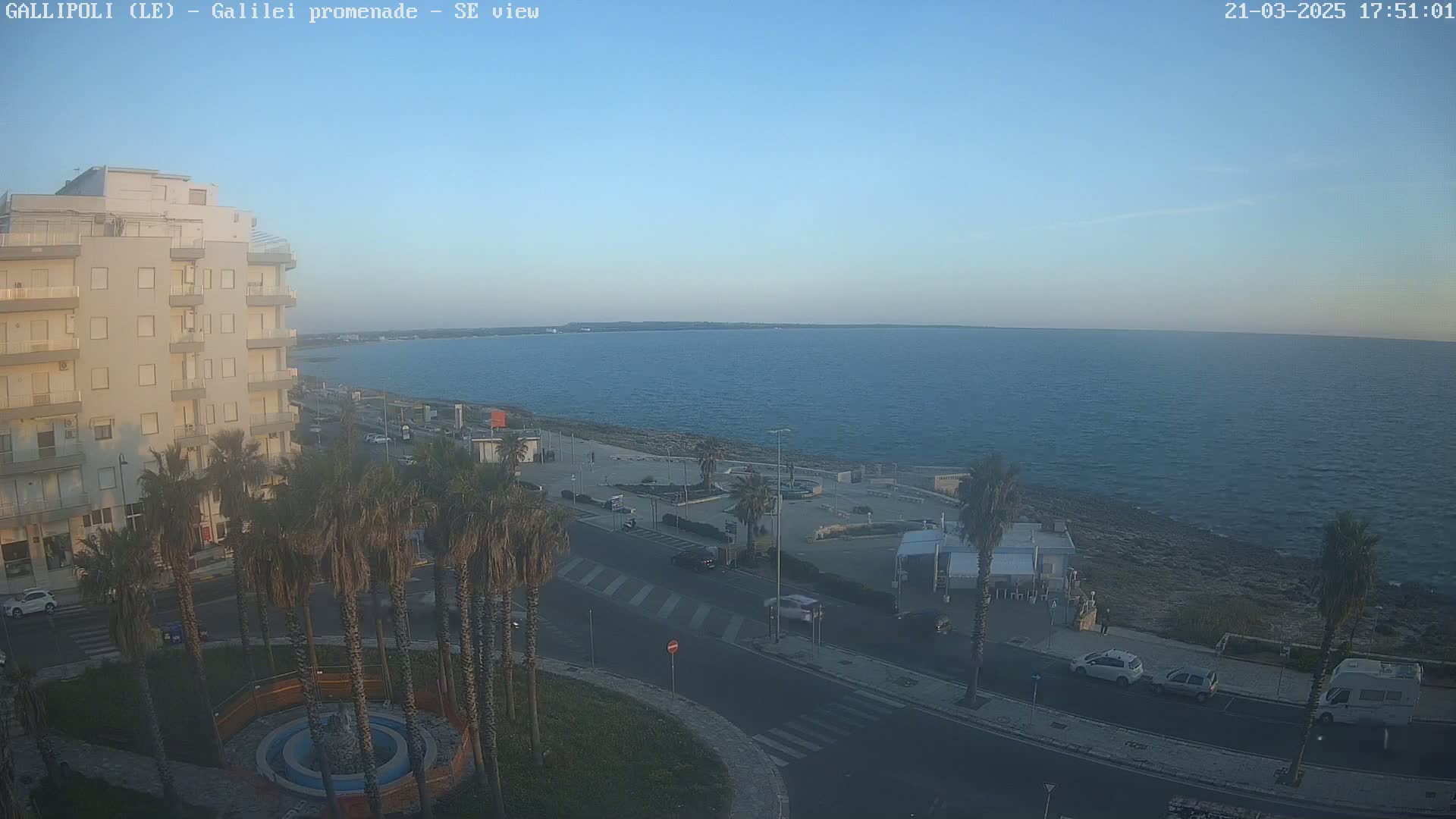 A seaside town scene at dusk shows a calm sea, a multi-story building, palm trees lining a road, and a few cars parked along the waterfront.