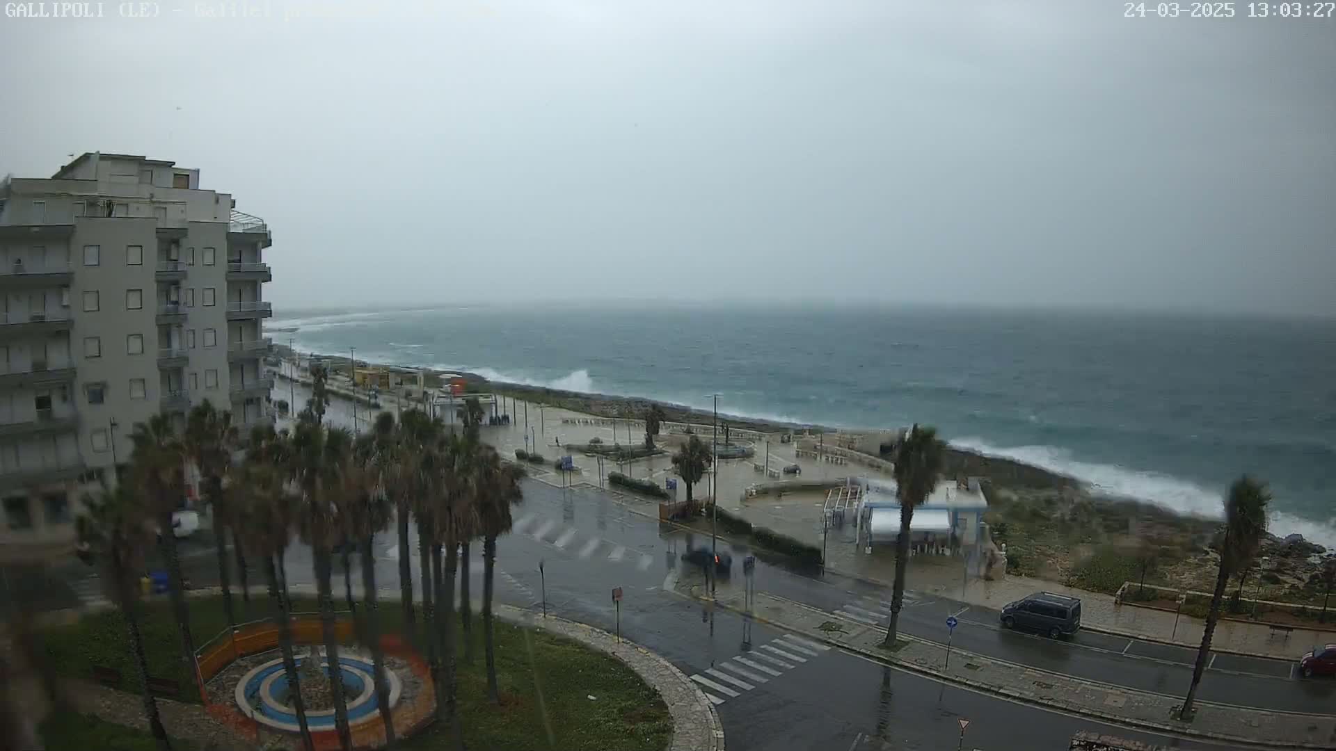 A multi-story building overlooks a rain-soaked coastal road and turbulent ocean waves under a gray, overcast sky.