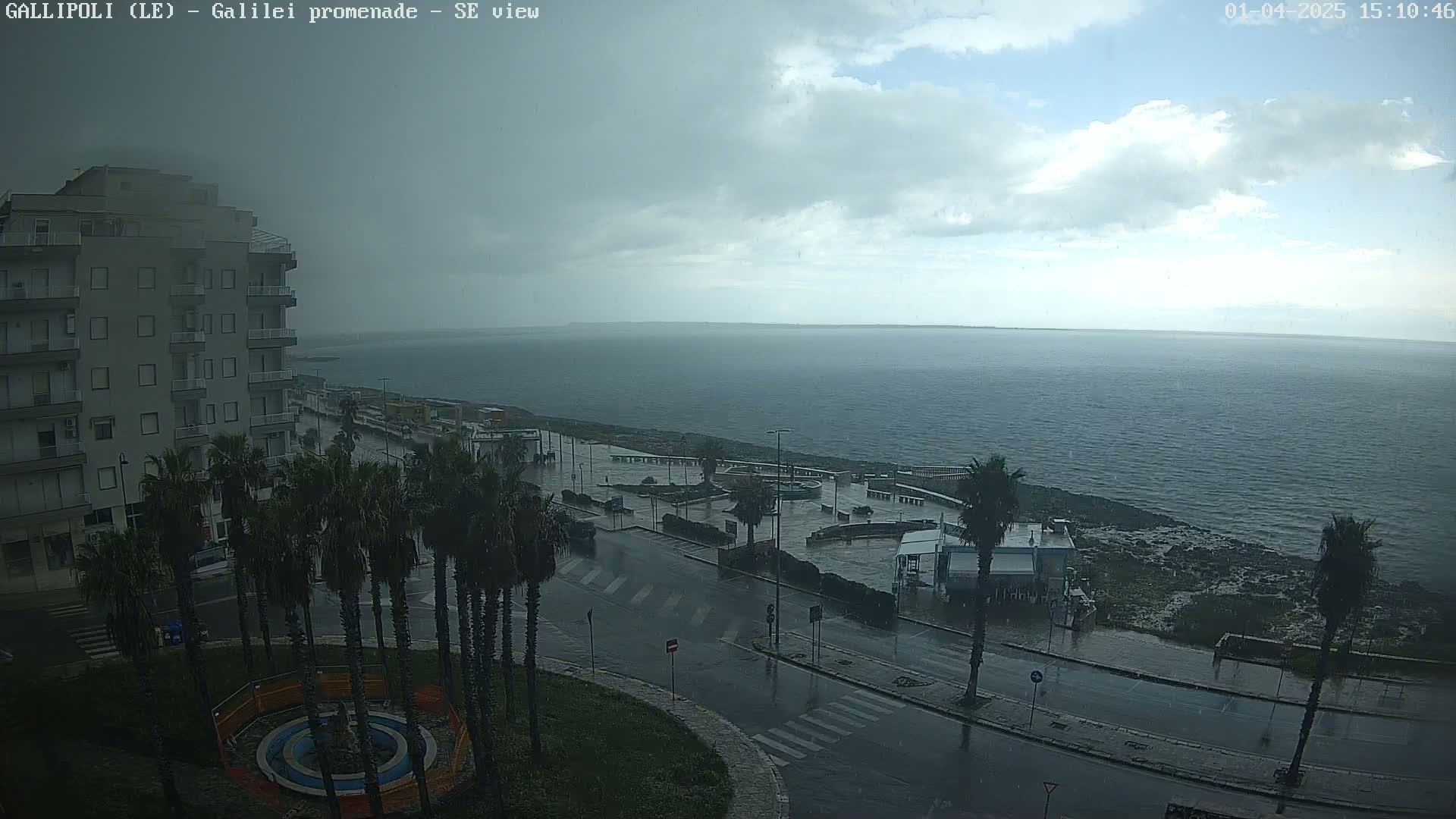 A seaside city street scene shows rain falling on palm trees, buildings, and wet roads leading to a rocky shore and a calm sea under a cloudy sky.