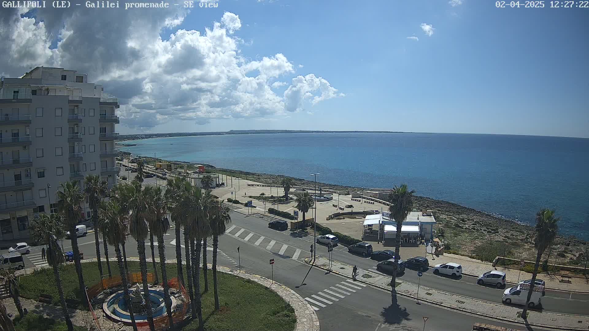A partly sunny day reveals a coastal town scene with a roadway, palm trees, a building, and a calm ocean under a partly cloudy sky.