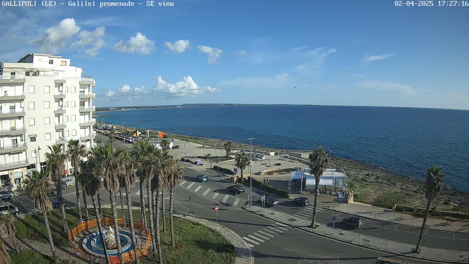 A seaside town scene under a mostly sunny sky shows a multi-story building, palm trees lining a road, and a calm blue sea stretching to the horizon.