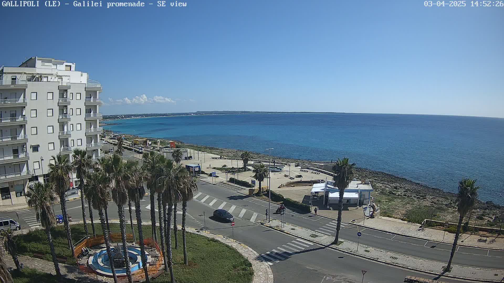 A seaside town with palm trees lining a road leading to a clear blue ocean under a mostly sunny sky.