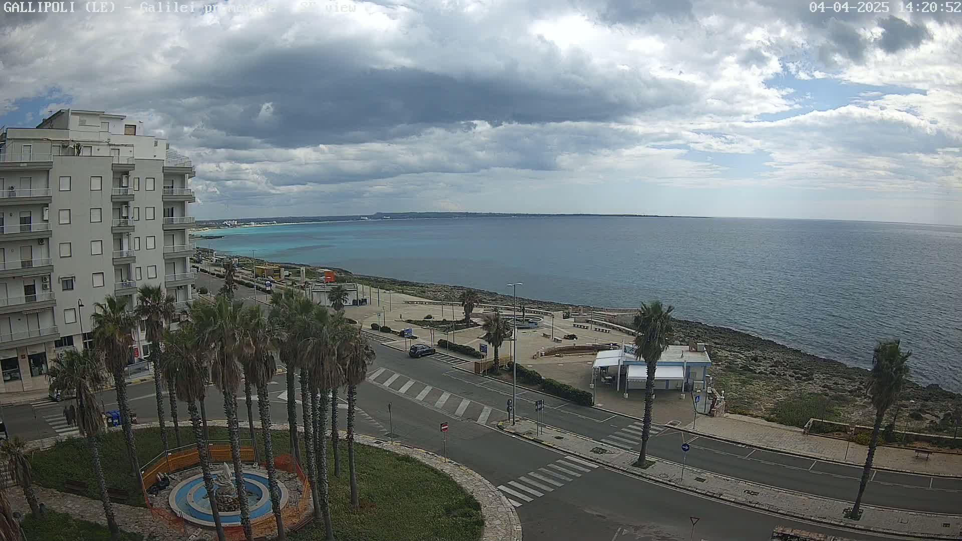 A partly cloudy day overlooks a coastal town with a calm sea, palm trees lining a street near a waterfront plaza.