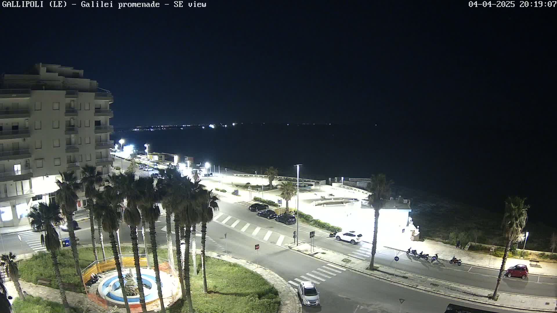 A nighttime, coastal scene shows a roadway lined with palm trees, a few parked cars and some motorcyclists, with a multi-story building and a calm dark sea in the background.