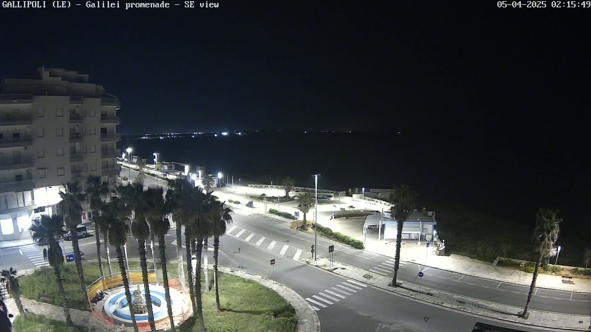 A nighttime, high-angle view of a coastal town's promenade, featuring a multi-story building, palm trees, and a calm, dark body of water in the background.