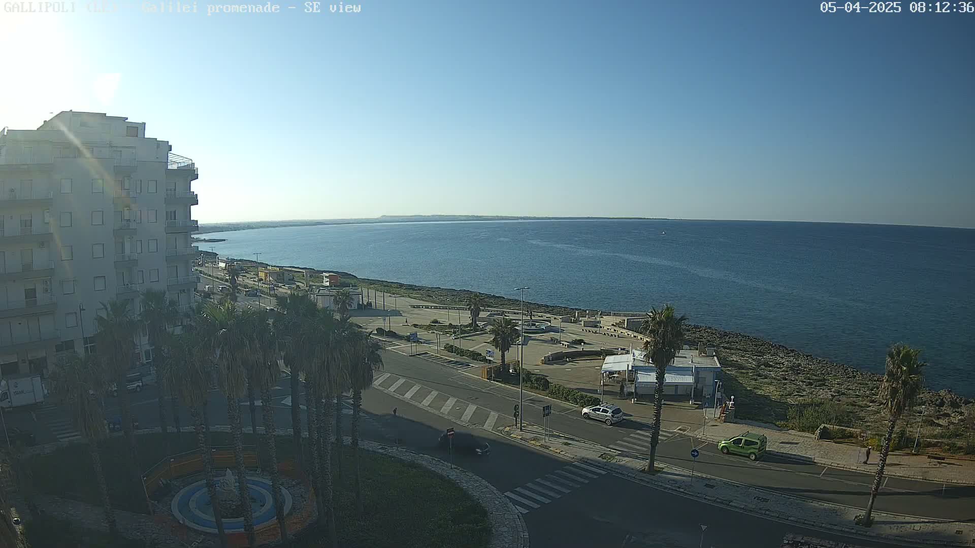A seaside town on a sunny day shows a calm ocean, a paved promenade, palm trees, and a multi-story building.