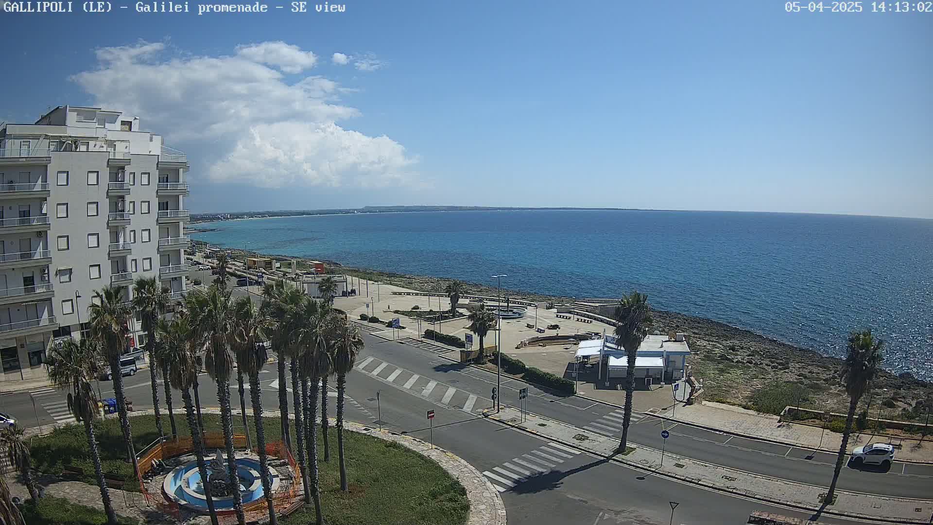 A partly cloudy day shows a seaside town with palm trees lining a street next to a calm blue ocean.