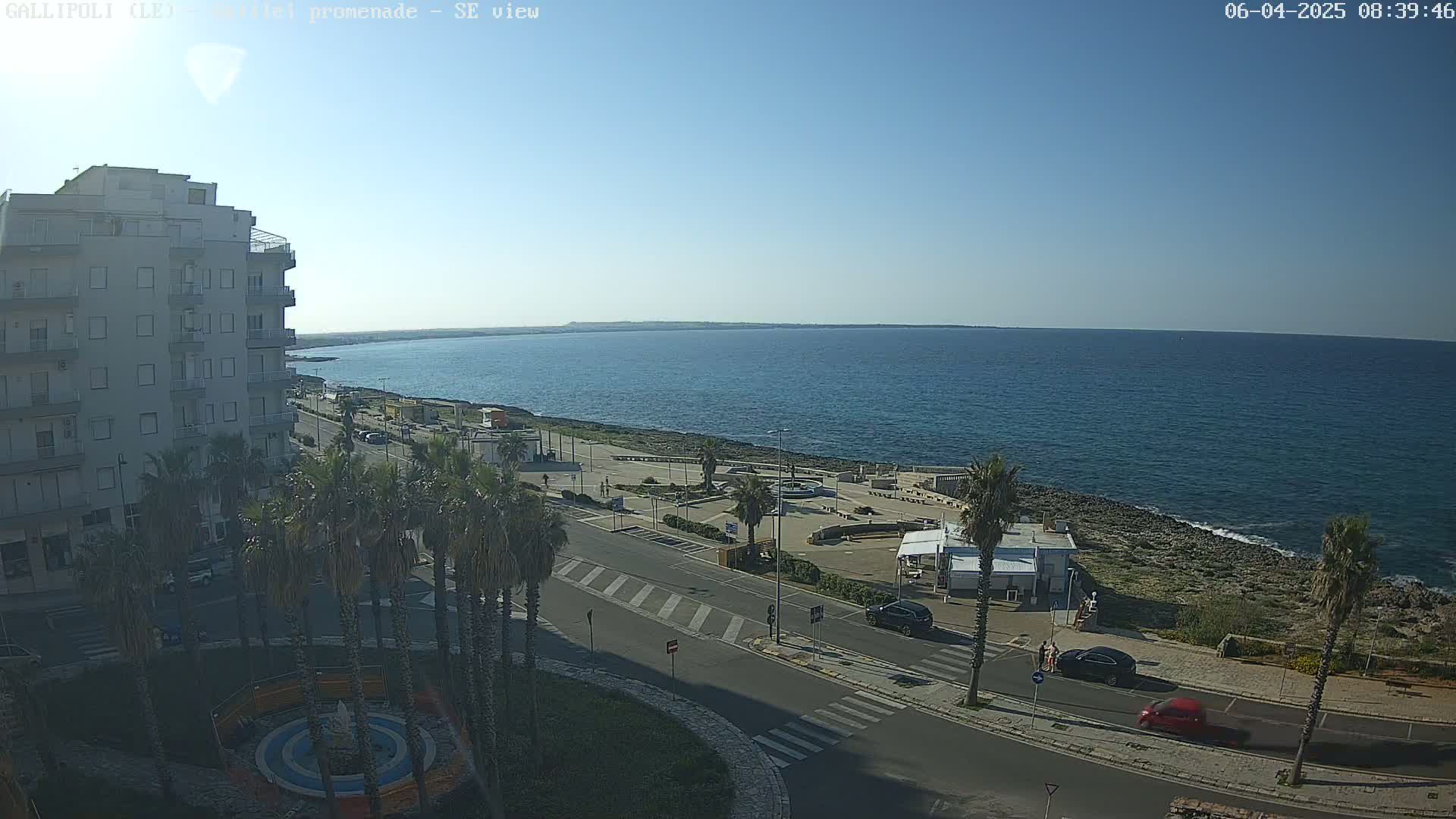 A seaside town on a clear, sunny day shows a multi-story building, palm trees, a paved road, a small plaza, and calm blue ocean waters.