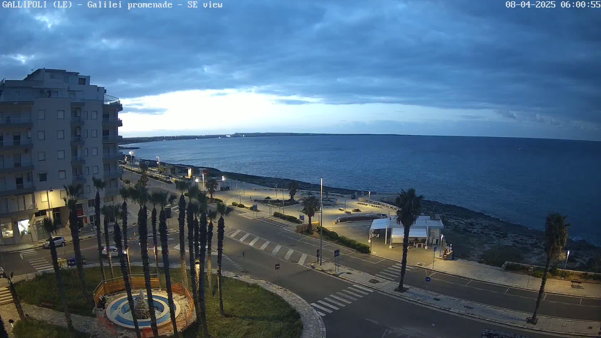 A seaside promenade at dusk, under a mostly cloudy sky, features palm trees lining a road that curves alongside a calm body of water.
