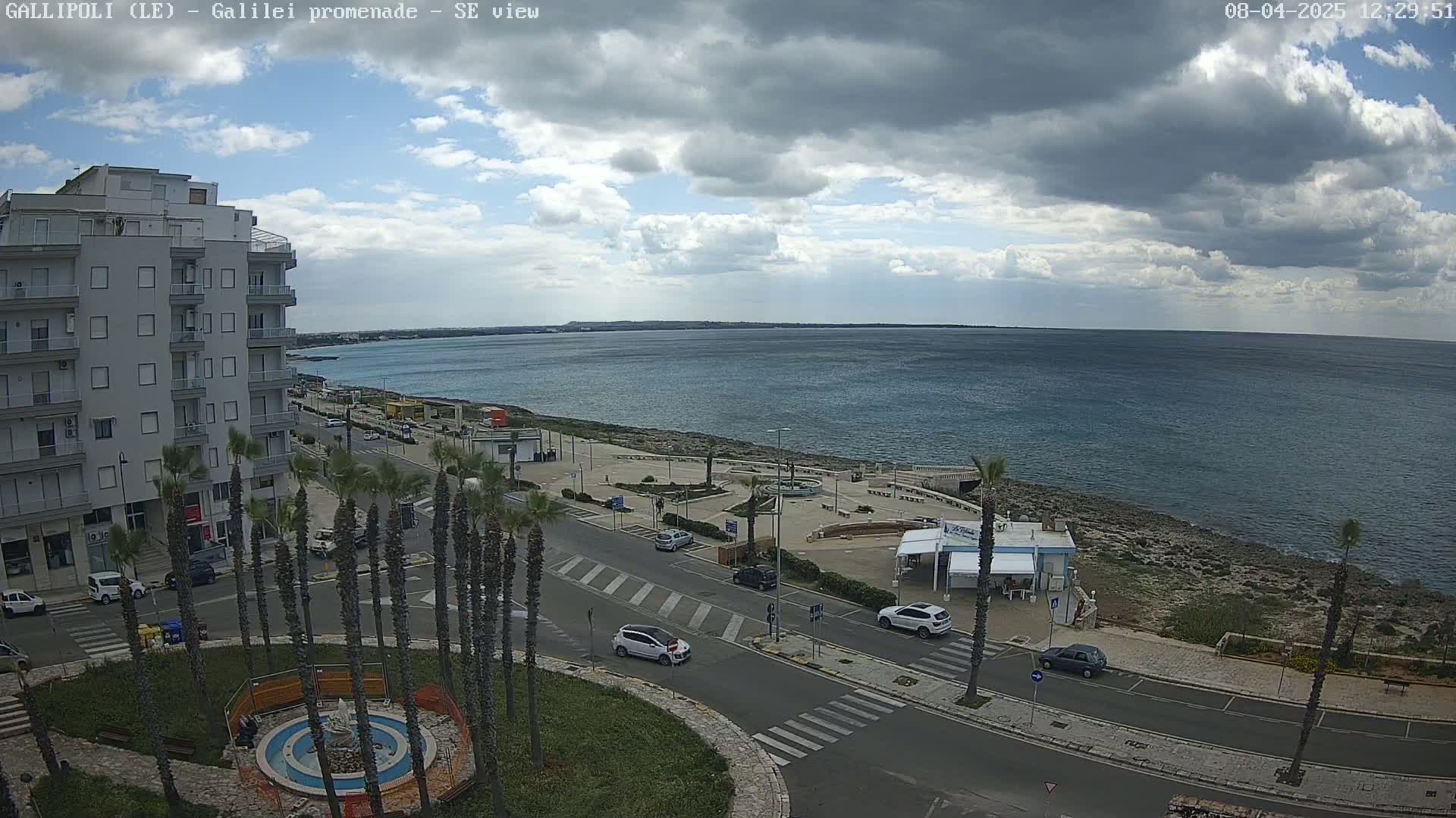 A seaside town scene under a partly cloudy sky shows a multi-story building, a coastal road with cars, palm trees, and a waterfront promenade with a calm sea beyond.