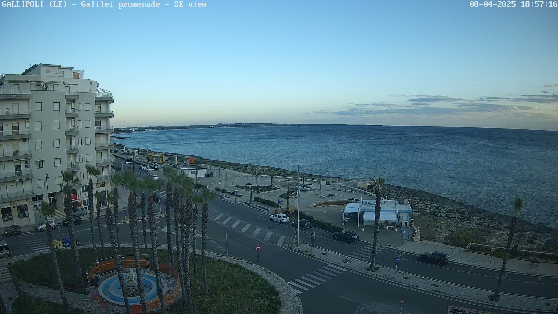 A seaside town scene under a clear, twilight sky shows a multi-story building, palm trees lining a road near a calm ocean, and a paved waterfront area.