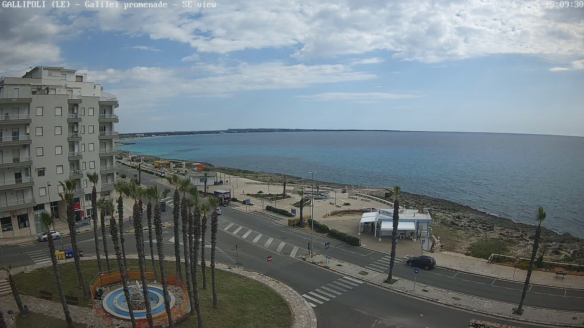 A partly cloudy day overlooking a coastal town with a promenade, palm trees, a multi-story building, and a calm blue sea.