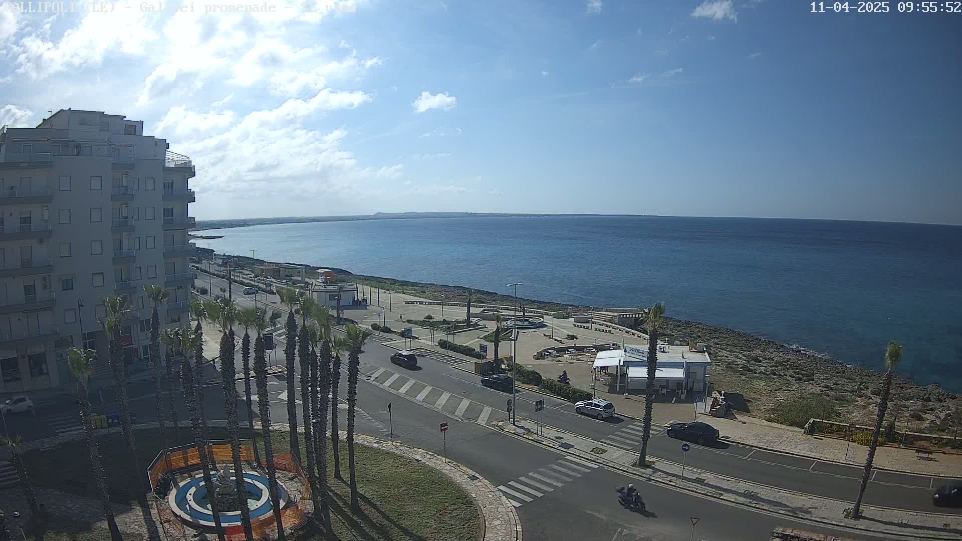 A partly cloudy day reveals a seaside town with a multi-story building, palm trees lining a road leading to a coastal promenade and a calm blue sea.