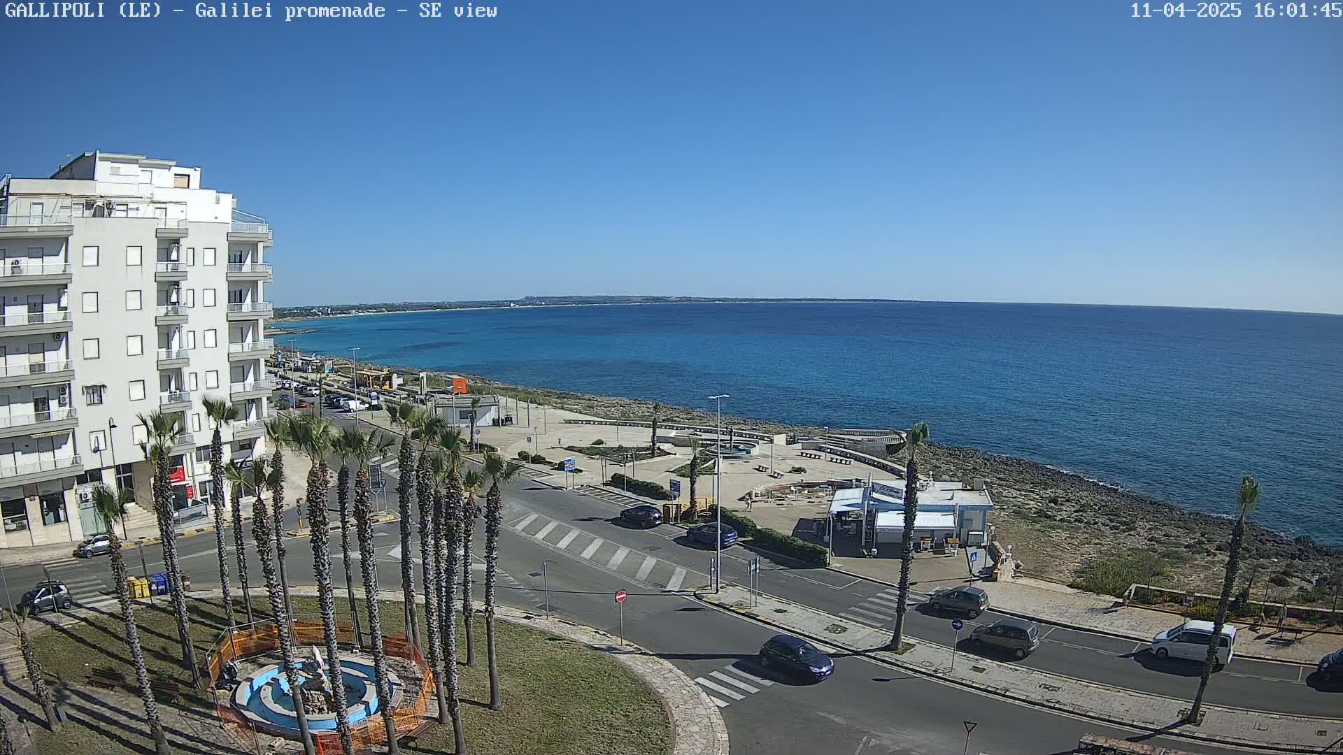 A sunny day overlooking a coastal town with a clear blue sea, palm trees lining a road near a waterfront promenade, and several cars parked along the streets.