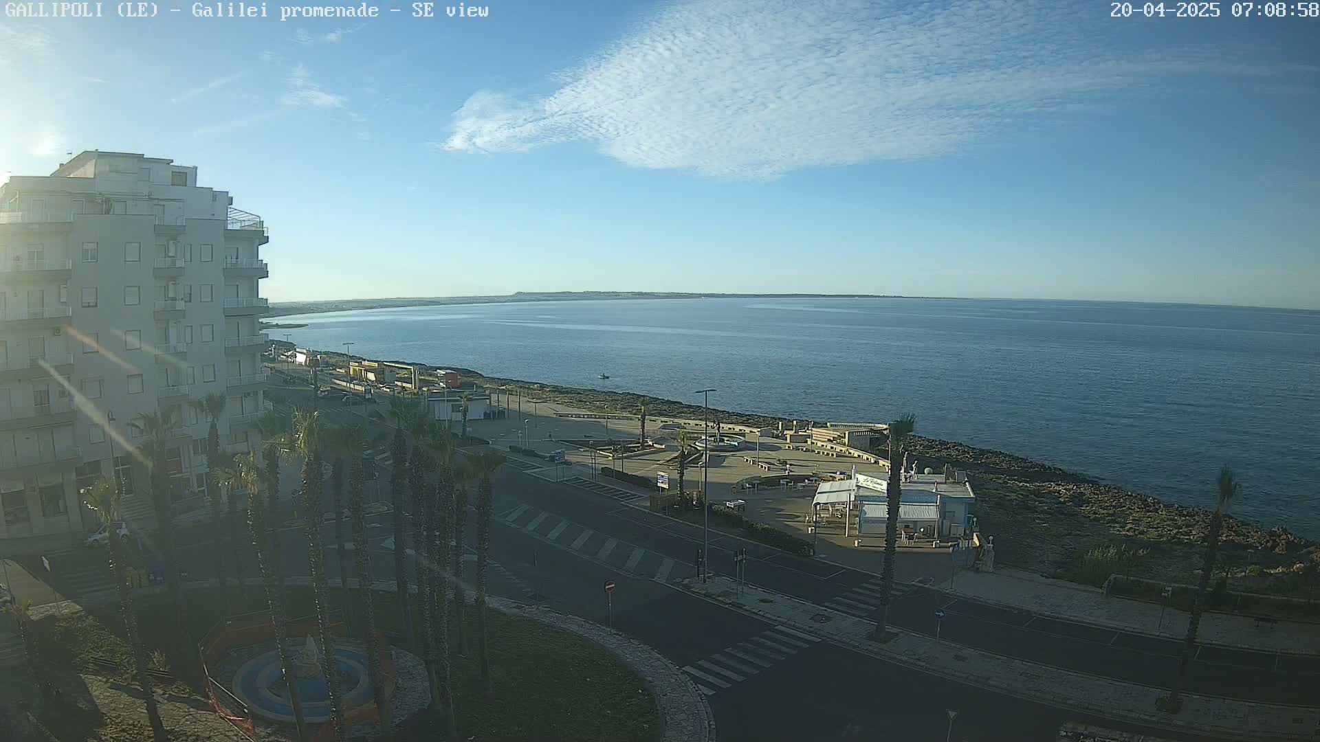 A mostly sunny day reveals a seaside promenade with palm trees, buildings, and a calm ocean extending to a distant shoreline.
