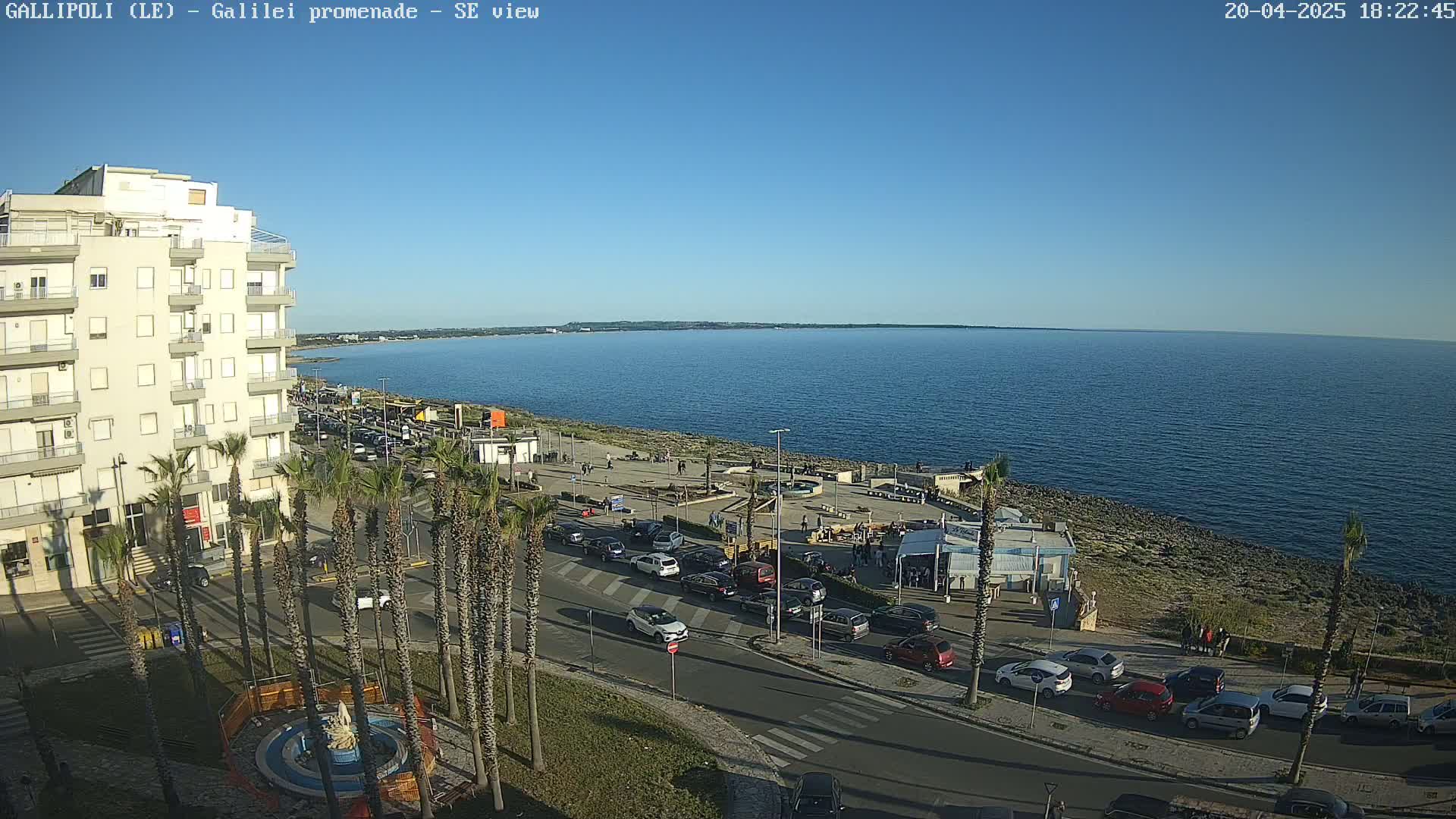 A seaside town scene under a clear blue sky shows a multi-story building, a palm tree-lined promenade with parked cars, and a calm ocean extending to the horizon.