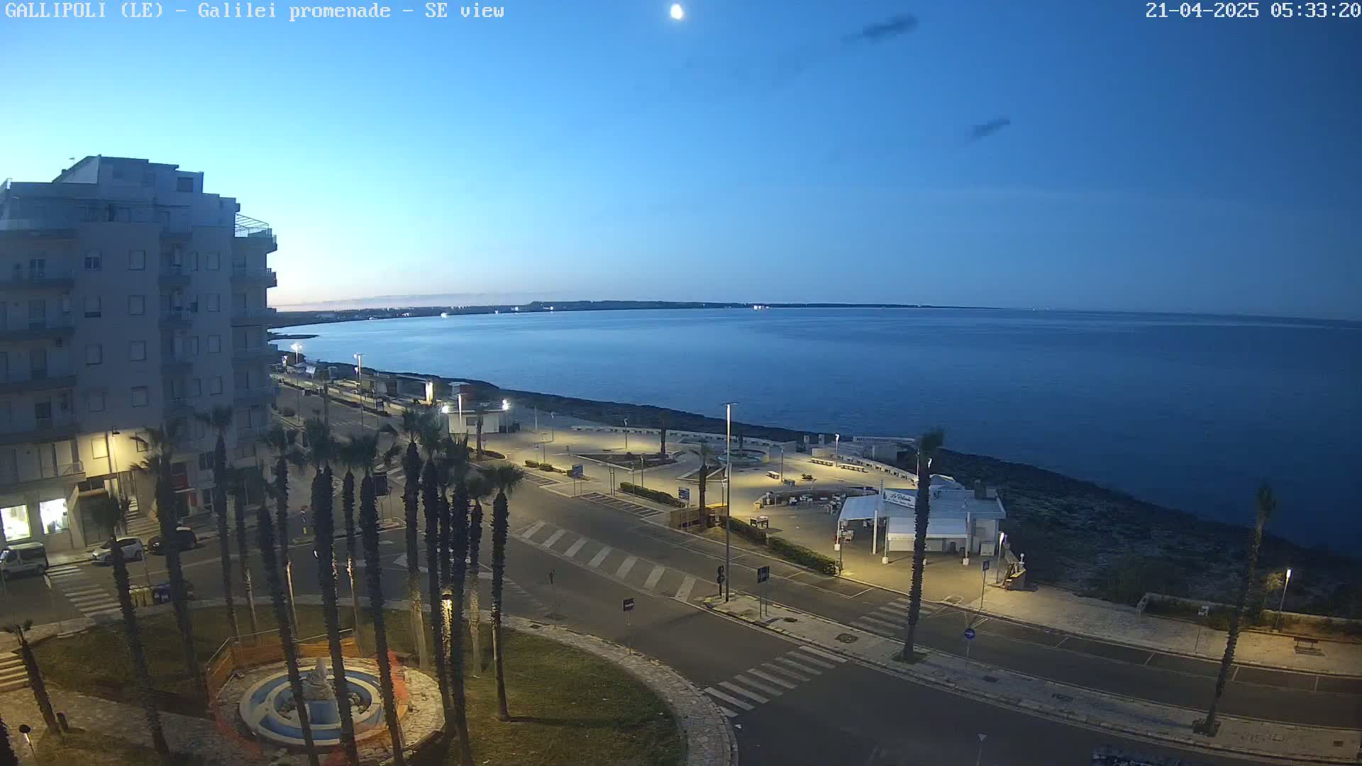 A seaside promenade at twilight, featuring a multi-story building, palm trees, and a calm, dark-blue sea under a clear sky.