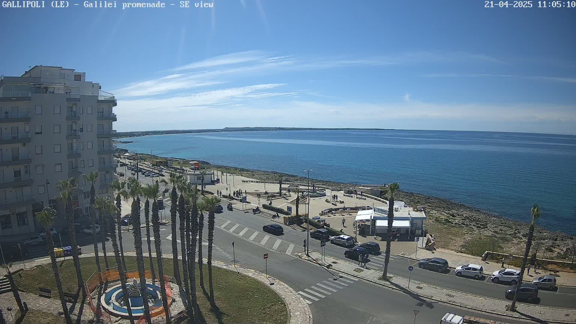 A sunny day shows a seaside town with a multi-story building, palm trees, a roadway, and a calm blue sea.