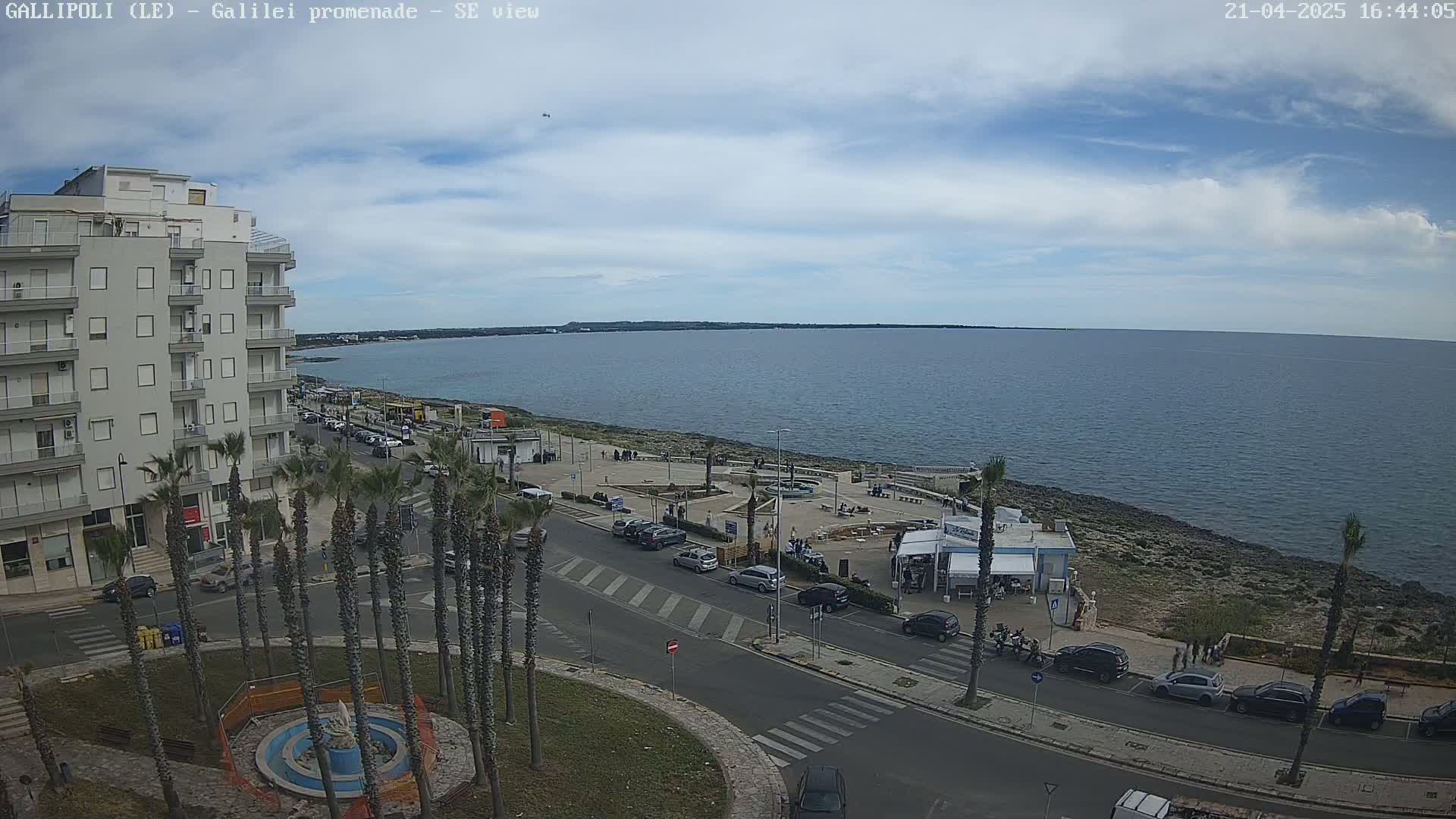 A partly cloudy day shows a seaside town with a multi-story building, palm trees, parked cars along a road near the ocean, and people gathered near a beachfront eatery.