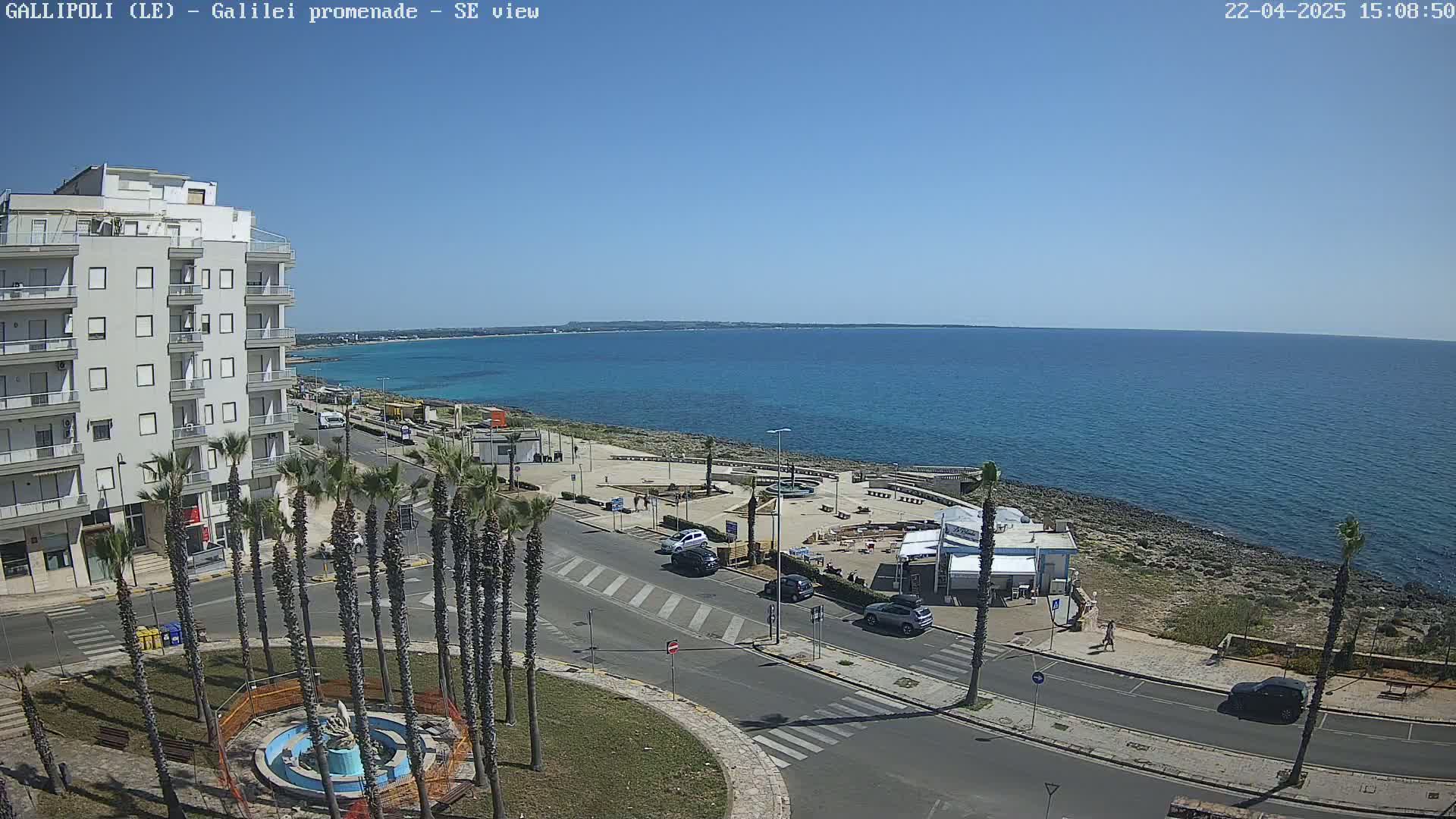 A seaside town on a sunny day shows a multi-story building, palm trees lining a road, and a calm blue sea extending to the horizon.