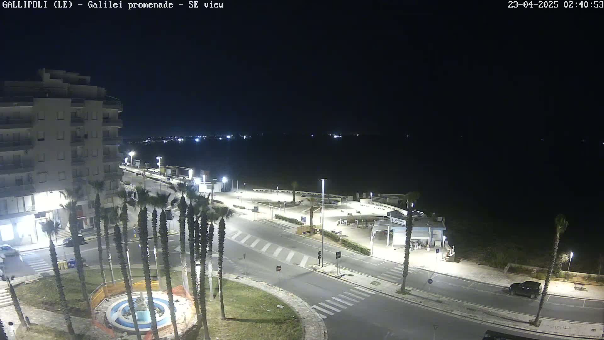 A nighttime, overhead view of a coastal town's promenade, featuring palm trees, buildings, and a calm sea.